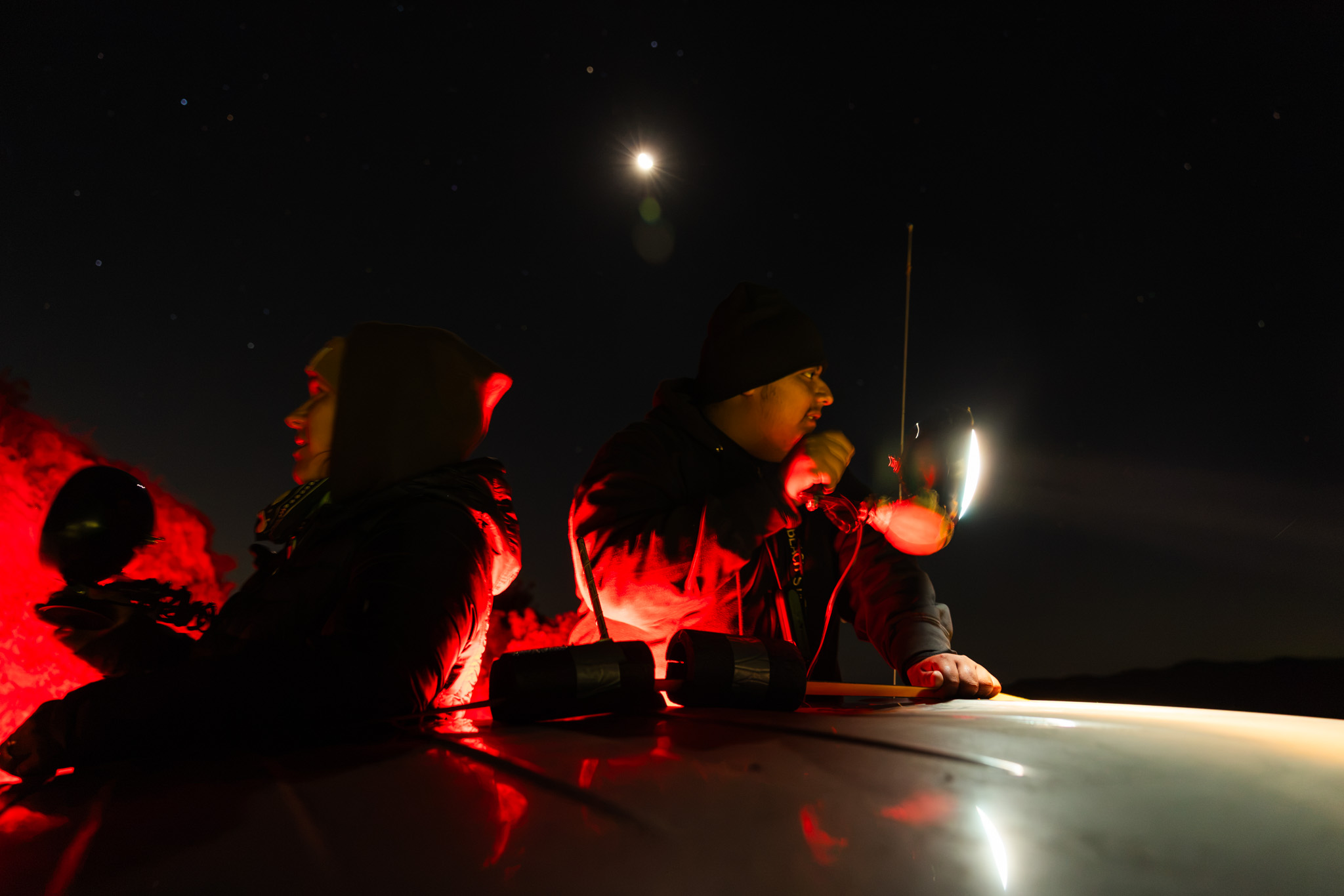 Two people stand behind a truck at night using red-lit headlamps and handheld lights under a starry sky